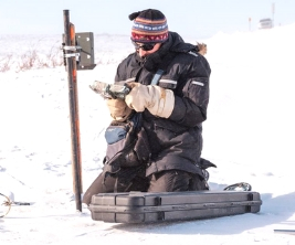 A researcher wearing a winter jacket, hat and mittens kneels in the snow in Northern Canada to conduct a research study.