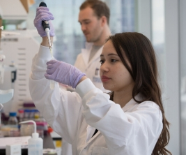 Une chercheuse portant des gants de sécurité et une blouse de laboratoire se tient dans un laboratoire et utilise un compte-gouttes pour déposer du liquide dans un petit récipient.