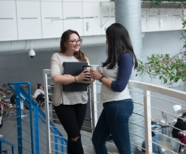 Two female students stand and talk on the second floor of a university building. 