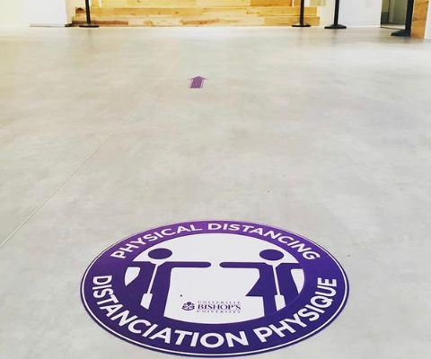 Un autocollant violet et blanc sur le sol d’un immeuble du campus de l’Université Bishop’s qui indique « PHYSICAL DISTANCING / DISTANCIATION PHYSIQUE ».