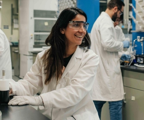 A female researcher wearing a white lab coat and safety goggles sits smiling in a university laboratory.